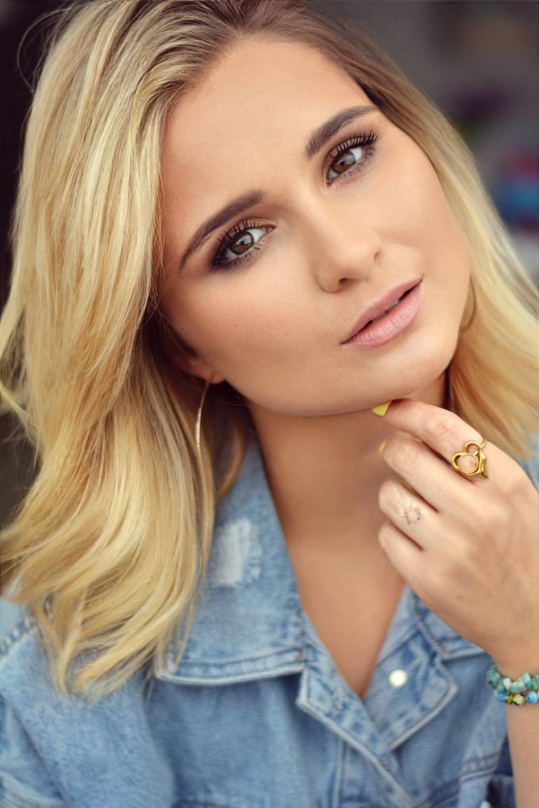 Portrait of a woman with blonde hair wearing a denim shirt, gold ring, and beaded bracelet, looking at camera